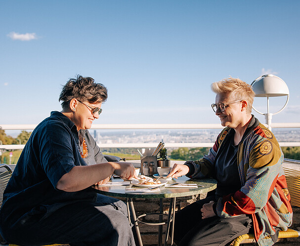 Zwei Frauen sitzen auf einer Terrasse und teilen sich einen Kuchen. Im Hintergrund Skyline von Wien. Mitglied im Expat Club werden.