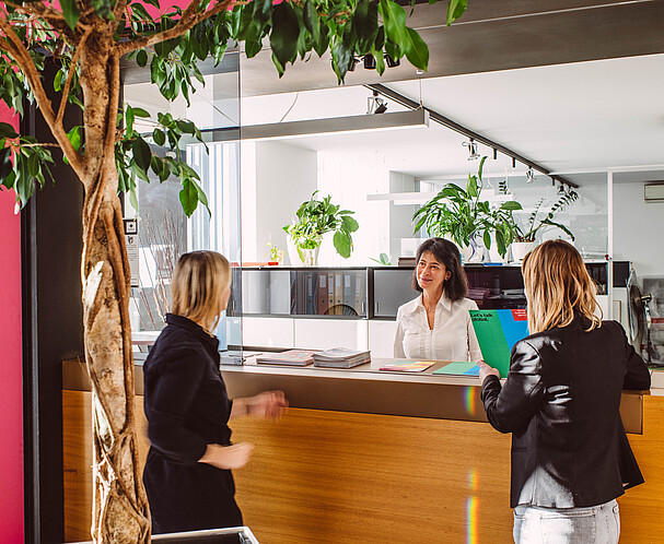 Zwei Frauen werden an einem hellen Empfangsbereich von einer anderen Frau willkommen geheißen. Expat Center: Leben und Arbeiten in Wien.